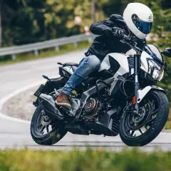 Motocyklista w białym kasku i czarnej kurtce pokonuje zakręt na drodze leśnej na motocyklu Bajaj Dominar 400.