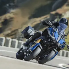 Motocyklista na niebieskim Yamaha Niken z bagażnikiem pokonuje zakręt na górskiej drodze.