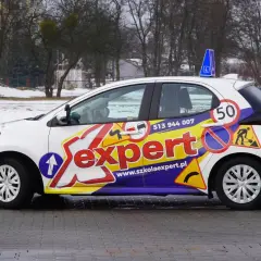Biały samochód szkoleniowy z napisem "Sexpert" i znakami drogowymi, gotowy do prowadzenia pojazdu.