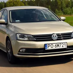 Złoty VW Jetta MK6 jedzie po drodze wśród zielonych pól i lasów.