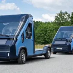 Dwa niebieskie, nowoczesne polskie samochody elektryczne jadące drogą pośród zieleni. Jeden to podwozie, drugi to furgonetka.