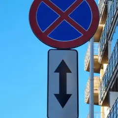 Znak zakaz zatrzymywania się z tabliczką wskazującą zakaz w obu kierunkach. Obok nowoczesny budynek z balkonami.