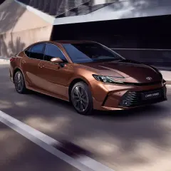 Nowa Toyota Camry Hybrid w kolorze miedzianym sunie po drodze, otoczona nowoczesną architekturą.