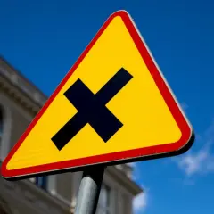 Żółty trójkąt z czarnym znakiem "X" ostrzega o skrzyżowaniu dróg. Uważaj, bo to droga z pierwszeństwem.