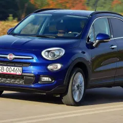 Niebieski Fiat 500 XL jedzie po drodze, otoczony jesiennym krajobrazem.