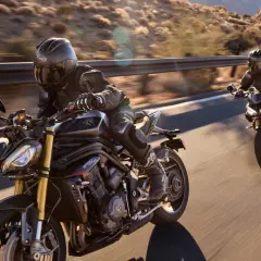 Dwóch motocyklistów na motocyklach Triumph Speed Triple pędzi po krętej drodze w słoneczny dzień.