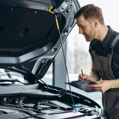 Mechanik w kombinezonie sprawdza silnik samochodu i notuje coś na tablicy.