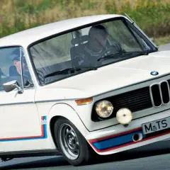 Białe BMW 2002 Turbo z kolorowymi pasami na błotnikach i zderzaku, z dwoma kierowcami w środku, pędzi po torze.