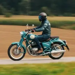 Mężczyzna w kasku i skórzanej kurtce jedzie turkusowym Royal Enfield Classic 650 po wiejskiej drodze, otoczonej polami.