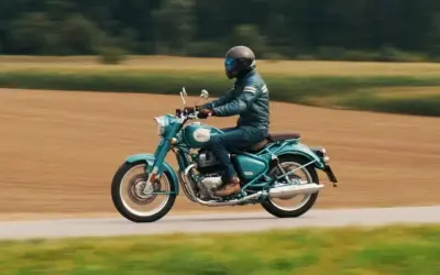 Mężczyzna w kasku i skórzanej kurtce jedzie turkusowym Royal Enfield Classic 650 po wiejskiej drodze, otoczonej polami.