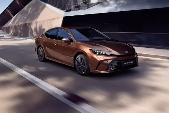 Nowa Toyota Camry Hybrid w kolorze miedzianym sunie po drodze, otoczona nowoczesną architekturą.