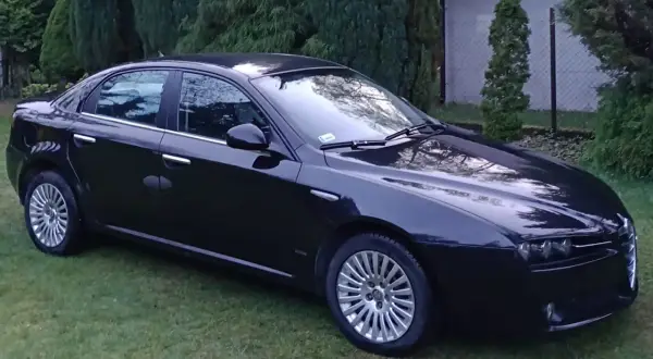 Elegancka, czarna Alfa Romeo 159 stoi na trawniku, otoczona zielenią.