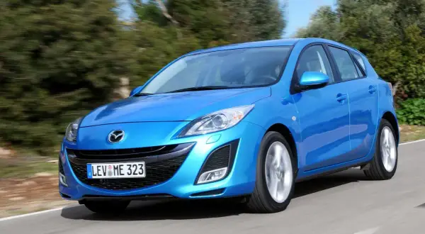 Niebieska Mazda 3 2010 w wersji hatchback pędzi po krętej drodze wśród zieleni.