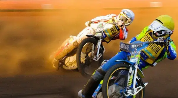 Dwóch zawodników ściga się na torze, ich motocykle żużlowe unoszą kurz.