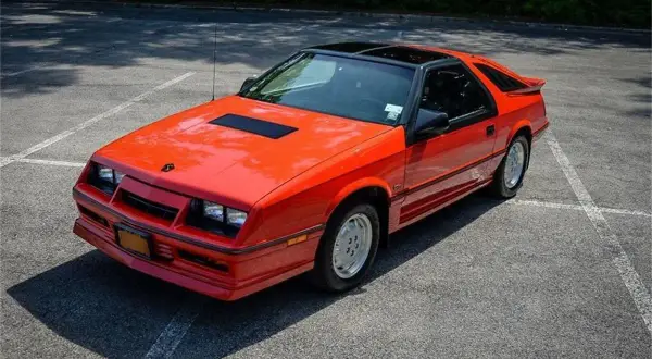 Pomarańczowy Dodge Daytona z czarnym dachem i spojlerem na parkingu.