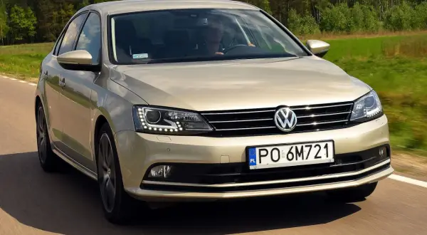 Złoty VW Jetta MK6 jedzie po drodze wśród zielonych pól i lasów.