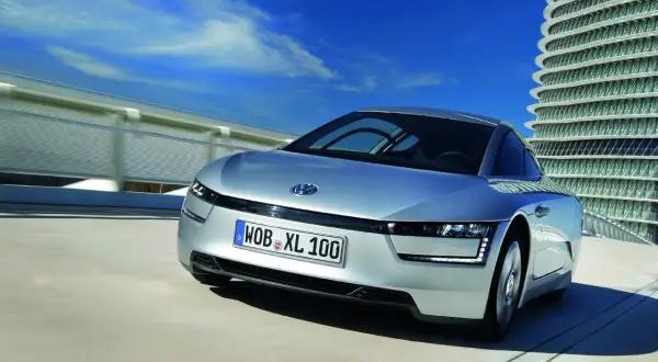 Srebrny Volkswagen XL1 sunie po drodze obok futurystycznego budynku.