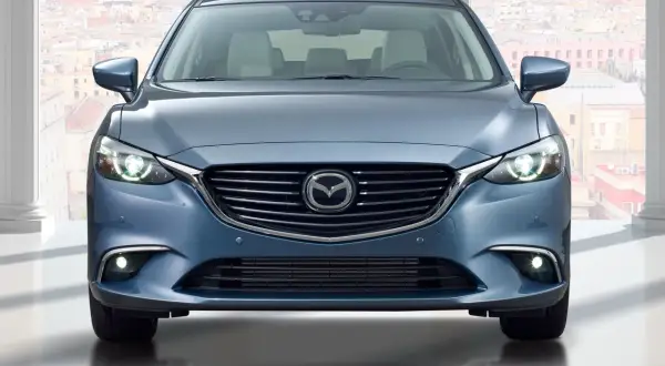 Niebieska Mazda 6 GJ kombi z przodu, z włączonymi światłami, na tle miasta.