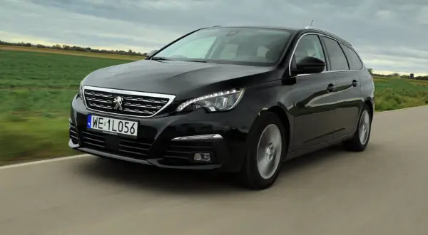 Czarny Peugeot 308 kombi z 2015 roku jedzie po drodze wśród zielonych pól.