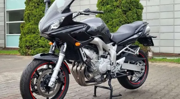 Czarny motocykl Yamaha FZ6, gotowy do jazdy, z czerwonymi akcentami na felgach, stoi przed budynkiem z zielonymi drzewami.