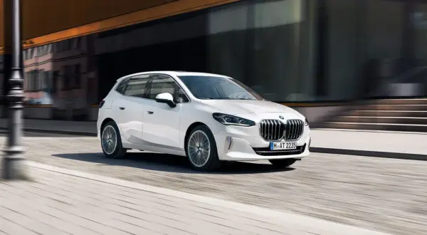 Białe BMW 218i sunie po miejskiej ulicy, odbijając światło od nowoczesnej fasady budynku.