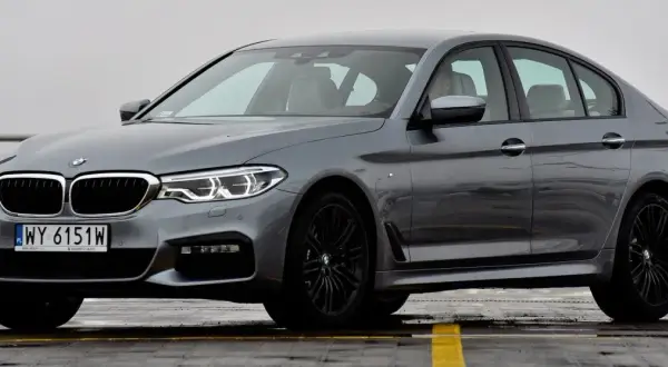 Szary sedan BMW Serii 5 G30 z czarnymi felgami, zaparkowany na mokrej nawierzchni.