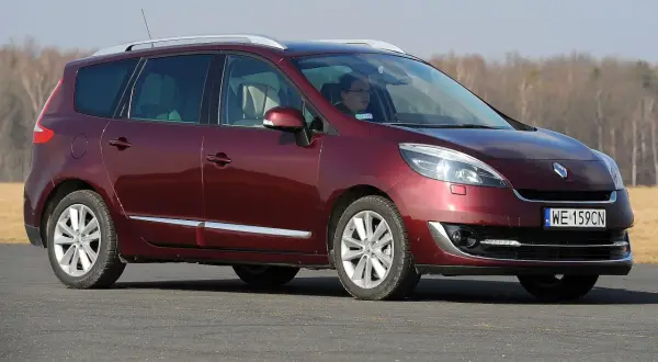 Ciemnoczerwony Renault Scenic 3 z kierowcą za kierownicą jedzie po drodze, w tle drzewa.