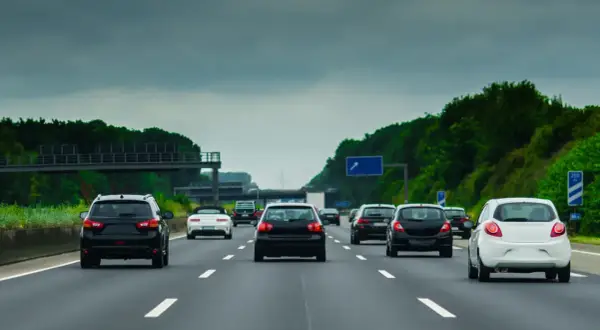 Samochody jadące drogą szybkiego ruchu pod pochmurnym niebem. W oddali widać zielone drzewa i wiadukt.
