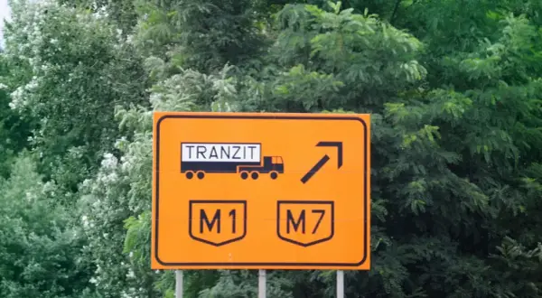 Pomarańczowy znak drogowy z napisem "TRANZIT" i symbolem ciężarówki, wskazujący zjazd na autostrady M1 i M7. Winieta węgry.