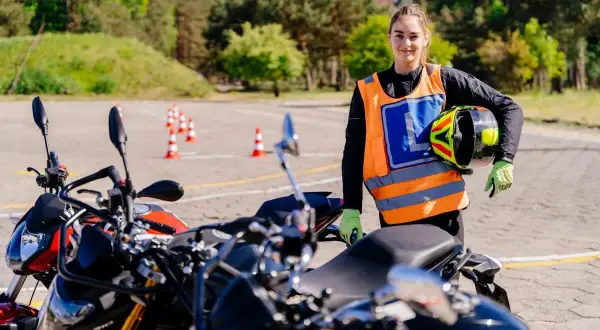 Młoda kobieta w kamizelce odblaskowej z literą L trzyma kask na placu manewrowym z motocyklami.
