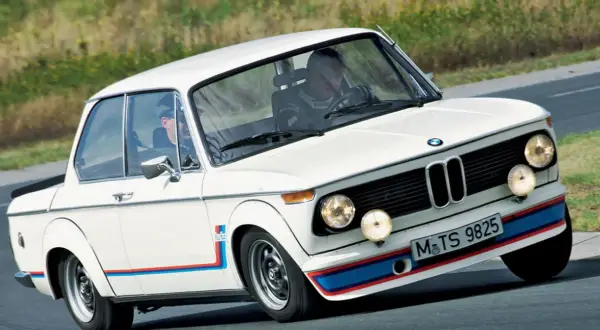 Białe BMW 2002 Turbo z kolorowymi pasami na błotnikach i zderzaku, z dwoma kierowcami w środku, pędzi po torze.