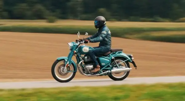 Mężczyzna w kasku i skórzanej kurtce jedzie turkusowym Royal Enfield Classic 650 po wiejskiej drodze, otoczonej polami.