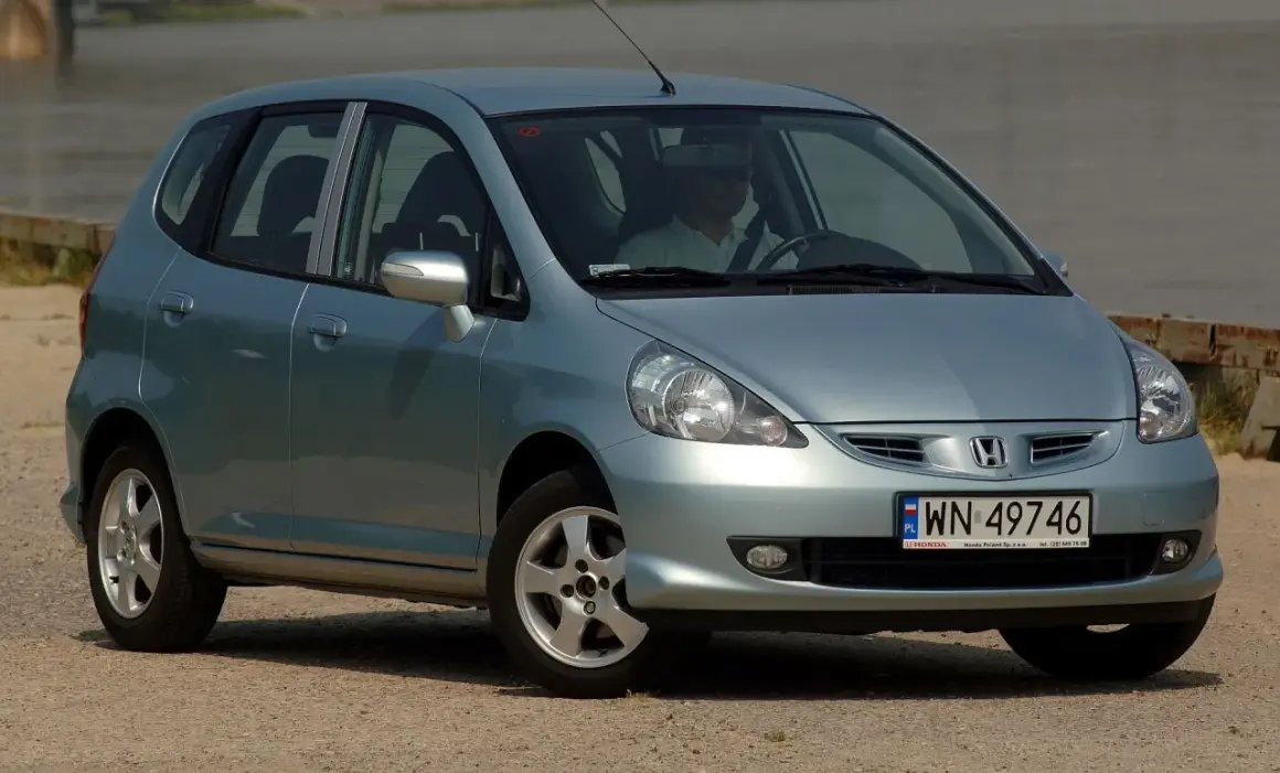 Jasnoszary Honda Jazz 2 z polskimi tablicami rejestracyjnymi WN 49746, zaparkowany przy wodzie.