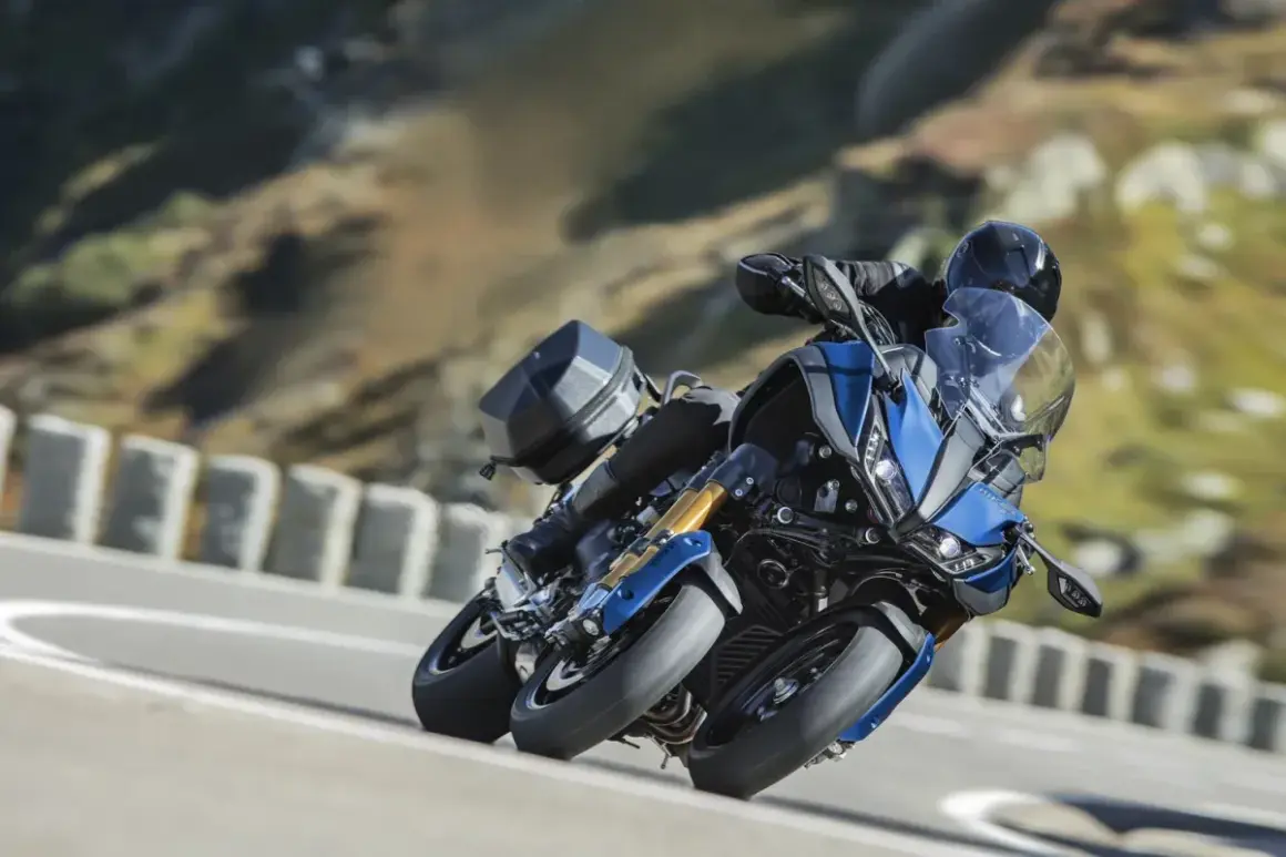 Motocyklista na niebieskim Yamaha Niken z bagażnikiem pokonuje zakręt na górskiej drodze.