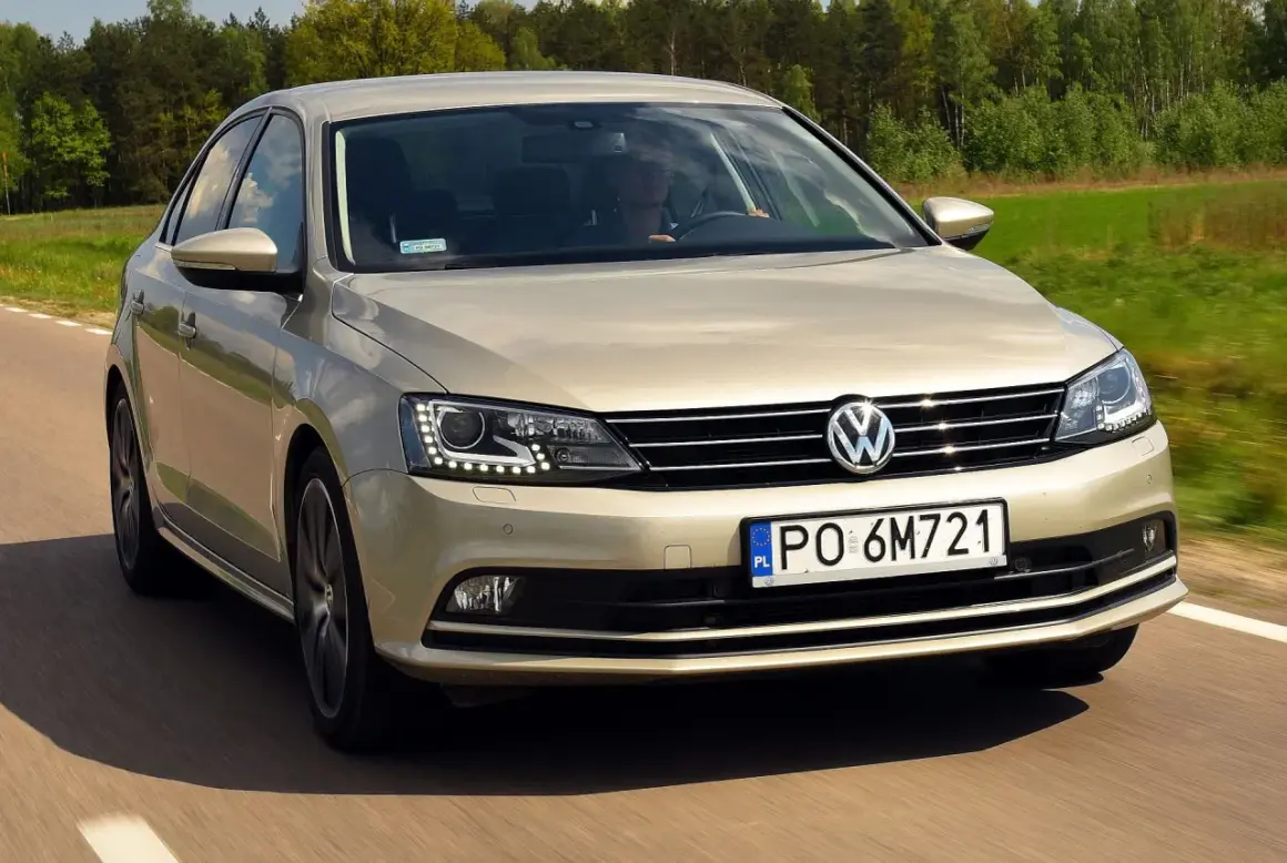 Złoty VW Jetta MK6 jedzie po drodze wśród zielonych pól i lasów.