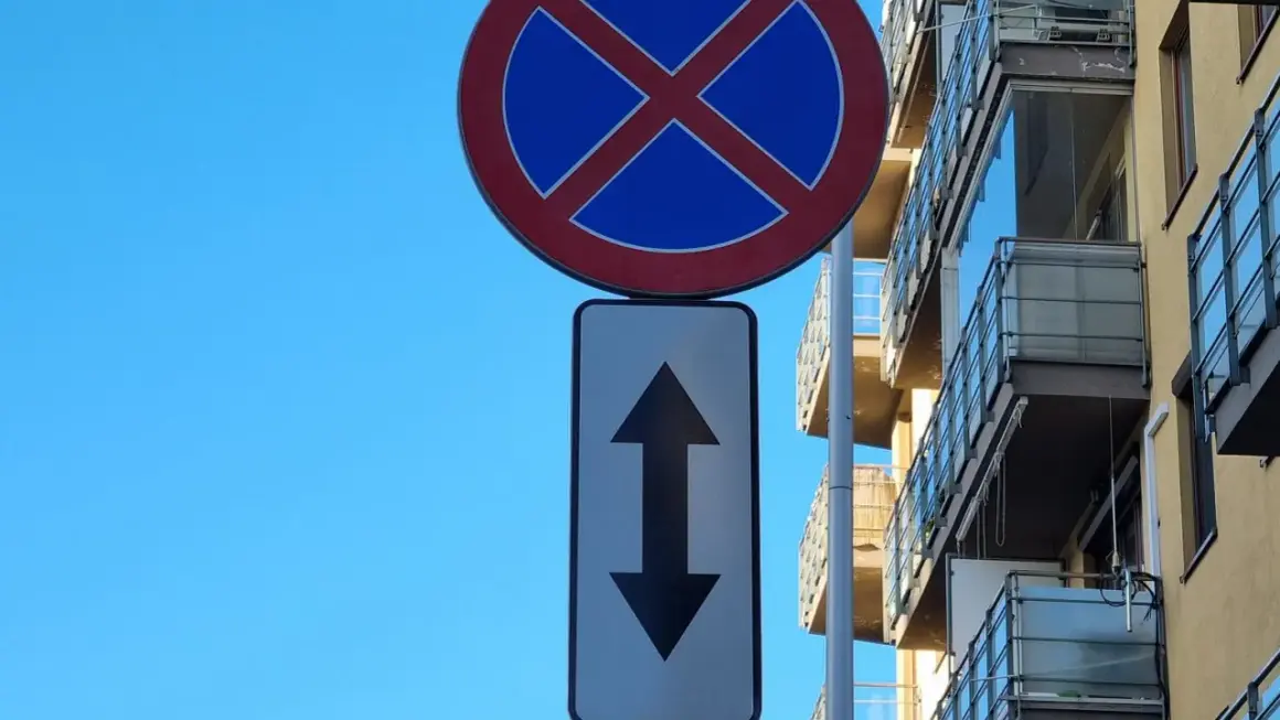 Znak zakaz zatrzymywania się z tabliczką wskazującą zakaz w obu kierunkach. Obok nowoczesny budynek z balkonami.