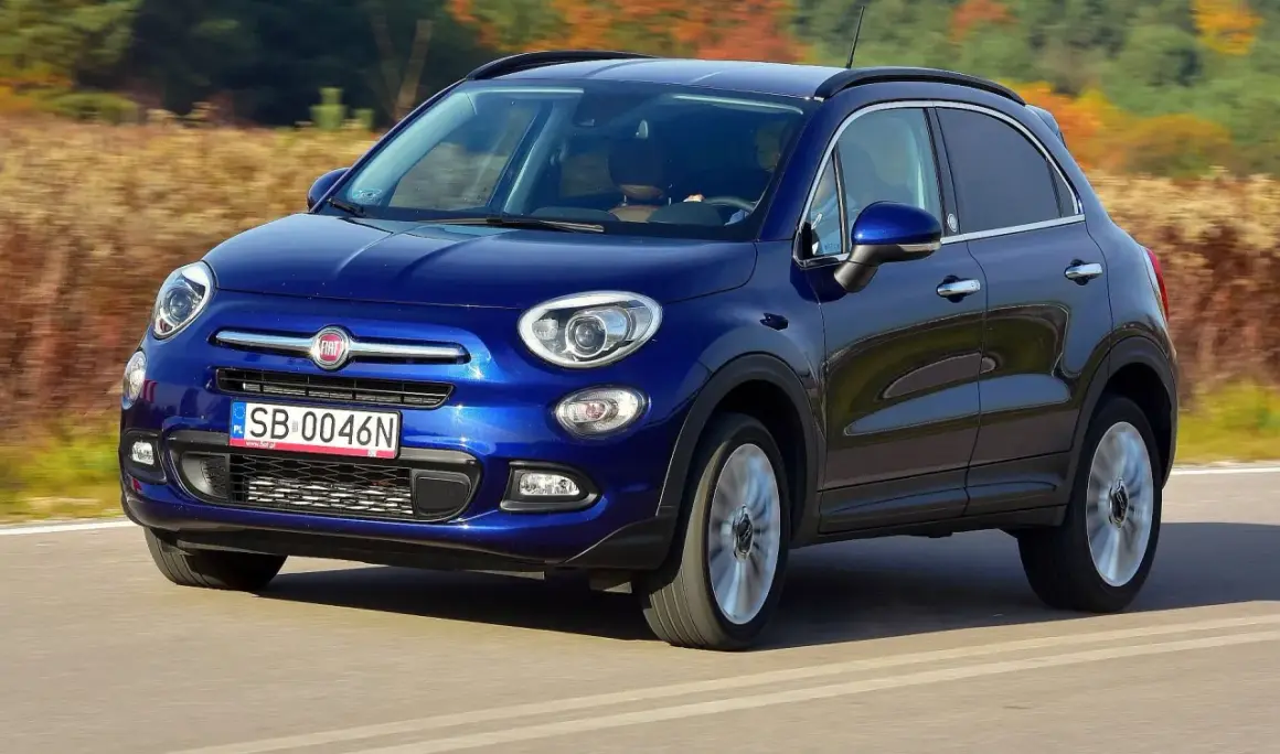 Niebieski Fiat 500 XL jedzie po drodze, otoczony jesiennym krajobrazem.