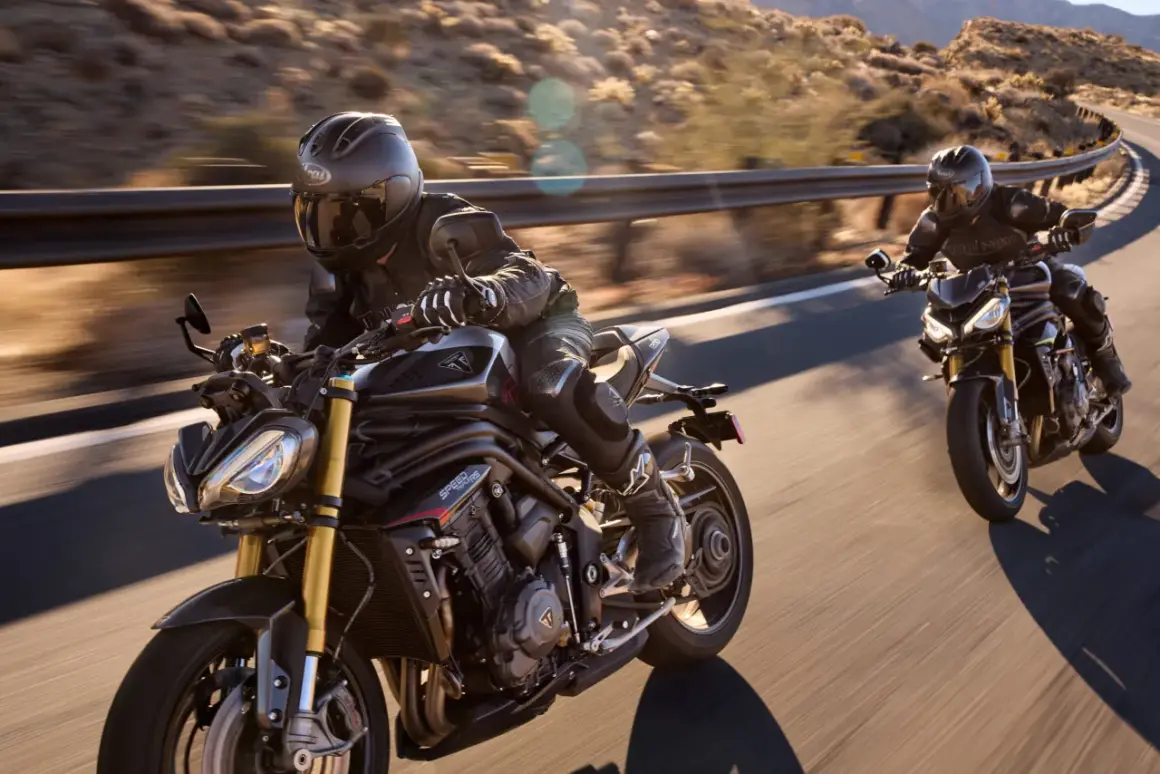 Dwóch motocyklistów na motocyklach Triumph Speed Triple pędzi po krętej drodze w słoneczny dzień.