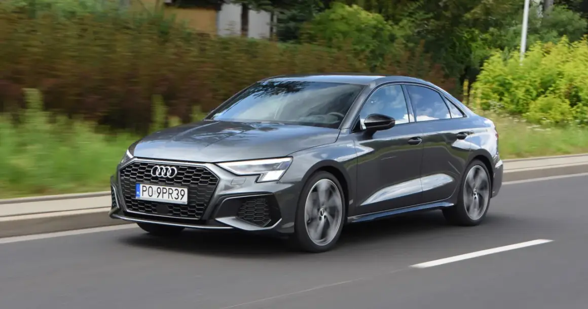 Szary Audi a3 sedan jedzie po drodze, otoczony zielenią.