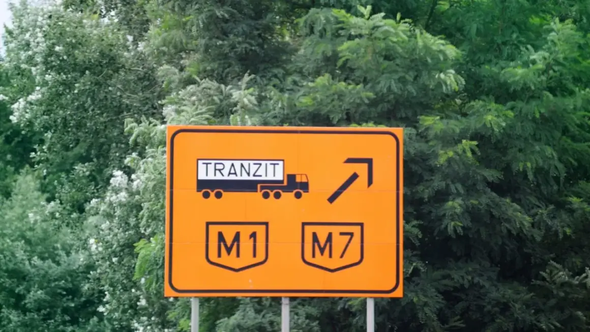 Pomarańczowy znak drogowy z napisem "TRANZIT" i symbolem ciężarówki, wskazujący zjazd na autostrady M1 i M7. Winieta węgry.