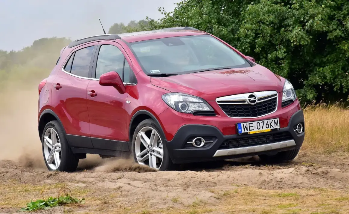 Czerwony Opel Mokka 2015 na piaszczystej drodze, unoszący kurz.