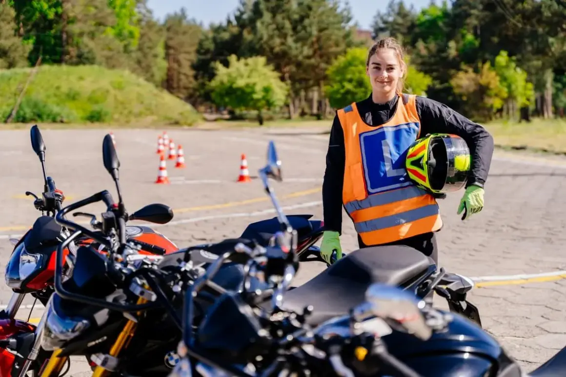 Młoda kobieta w kamizelce odblaskowej z literą L trzyma kask na placu manewrowym z motocyklami.
