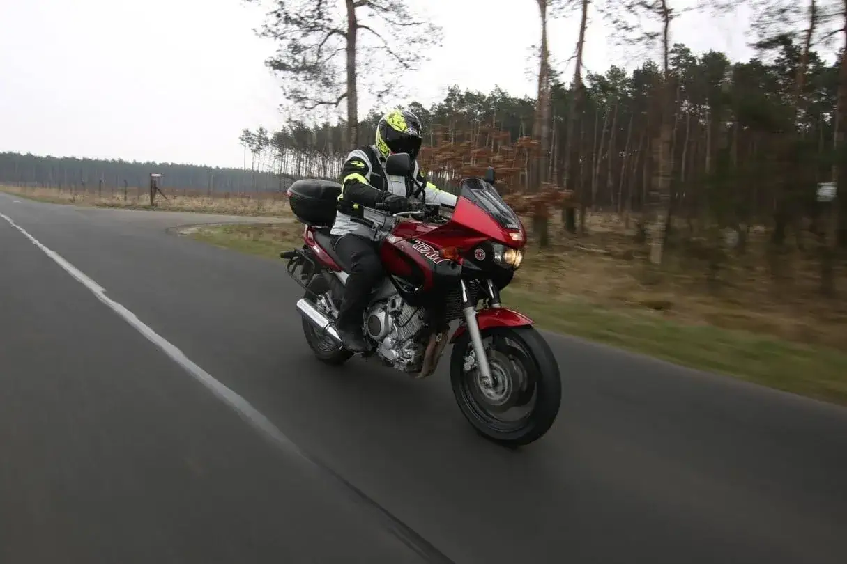 Motocyklista na czerwonym motocyklu Yamaha TDM 850 przemierza pustą drogę wśród drzew.