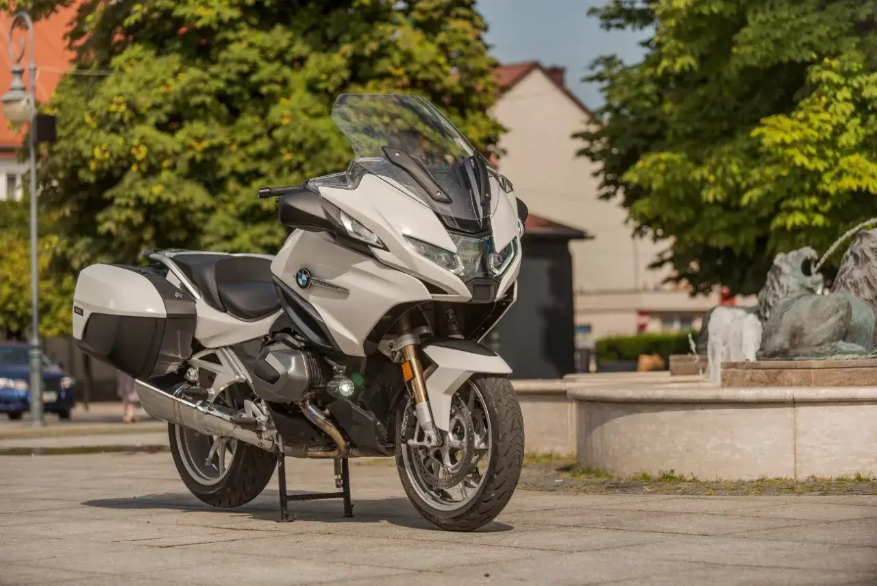 Biały motocykl BMW R 1250 RT z kuframi, zaparkowany na placu obok fontanny z lwami.