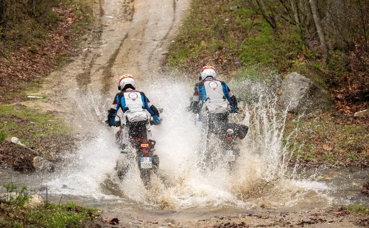 Dwóch motocyklistów na motocyklach Honda CB500X przemierza błotnistą kałużę, tworząc fontanny wody.