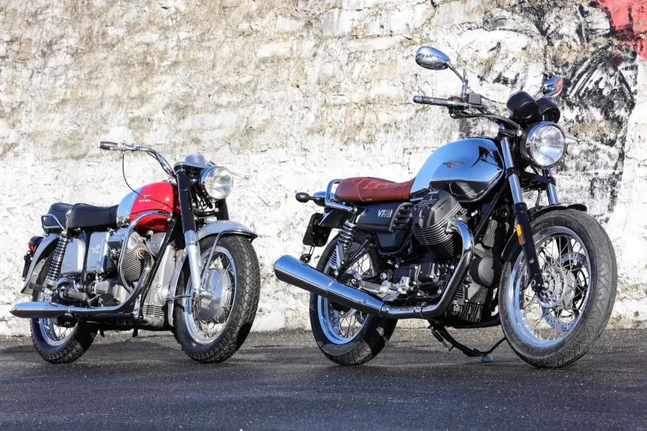 Dwa motocykle Moto Guzzi V7: jeden czerwono-srebrny, drugi chromowany z brązową kanapą, stoją przed ścianą z graffiti.