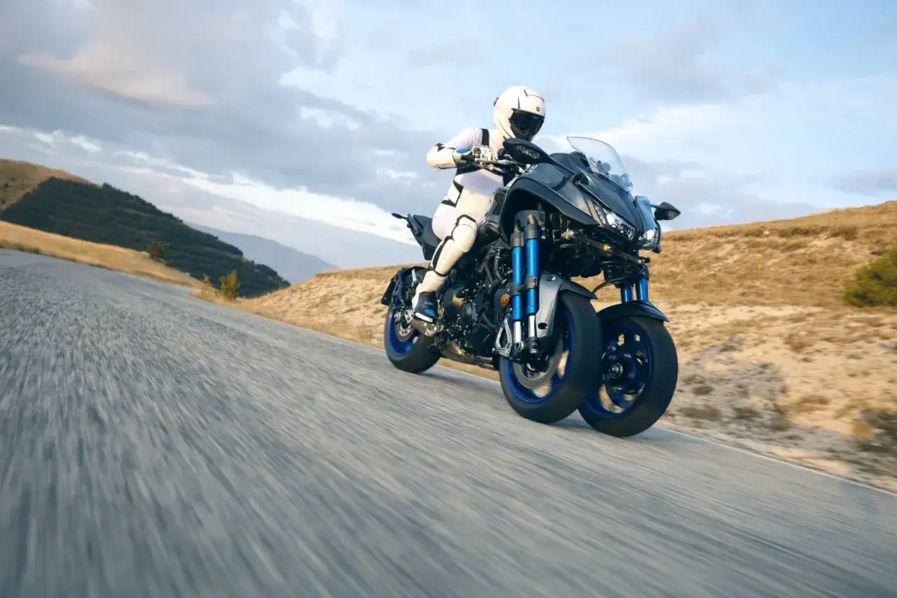 Motocyklista w białym kombinezonie i kasku pędzi na trójkołowym motocyklu Yamaha Niken po krętej drodze wśród pagórków.