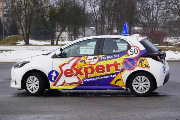 Biały samochód szkoleniowy z napisem "Sexpert" i znakami drogowymi, gotowy do prowadzenia pojazdu.