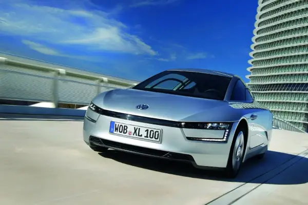 Srebrny Volkswagen XL1 sunie po drodze obok futurystycznego budynku.