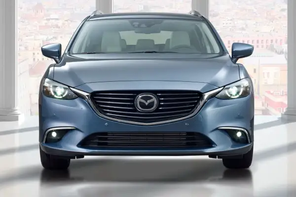 Niebieska Mazda 6 GJ kombi z przodu, z włączonymi światłami, na tle miasta.